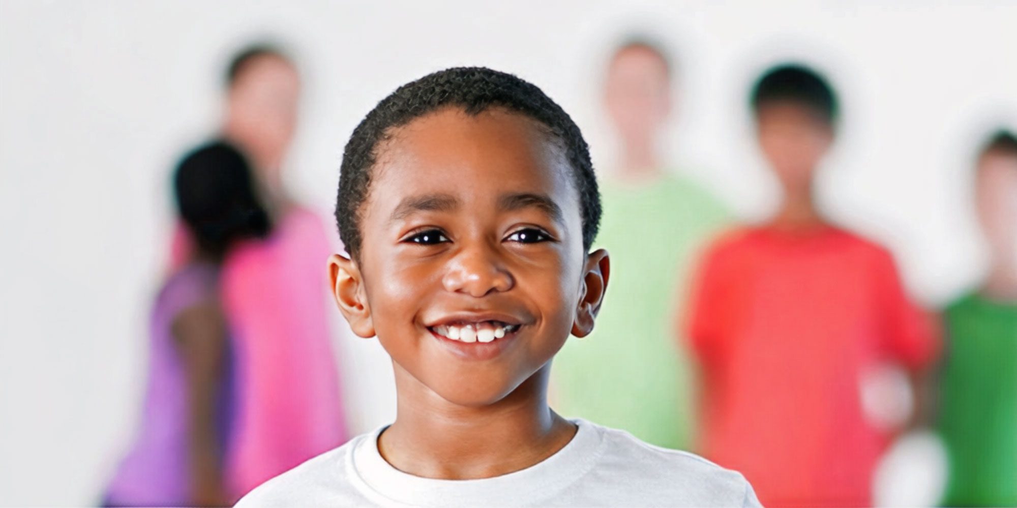 Smiling boy in a white T-shirt with classmates blurred behind him.